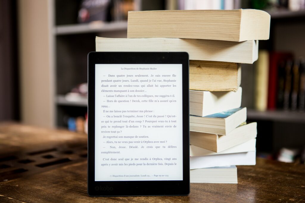 a black ereader leans against a stack of books. the ereader displays a page from a book.