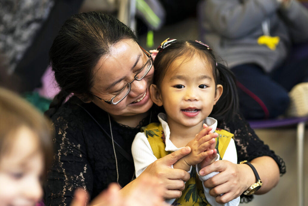 A young girl in a yellow dress with pigtails sits in her mum's lap, clapping and smiling.