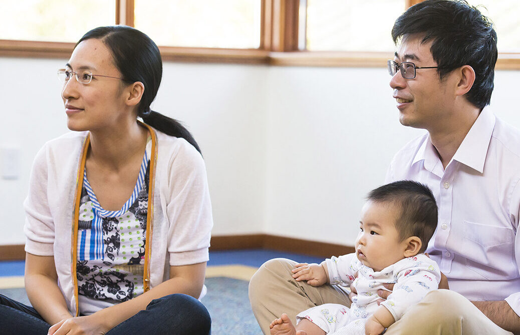 An infant sits on their caregivers lap, listening to a story. Another adult sits beside them and watches a storyteller out of frame.
