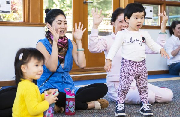 A group of adults and small children dance, clap, and sing.