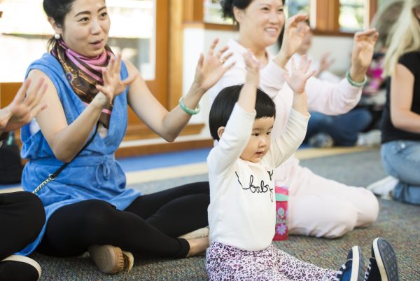 A group of adults and toddlers raise their arms, clapping and singing.