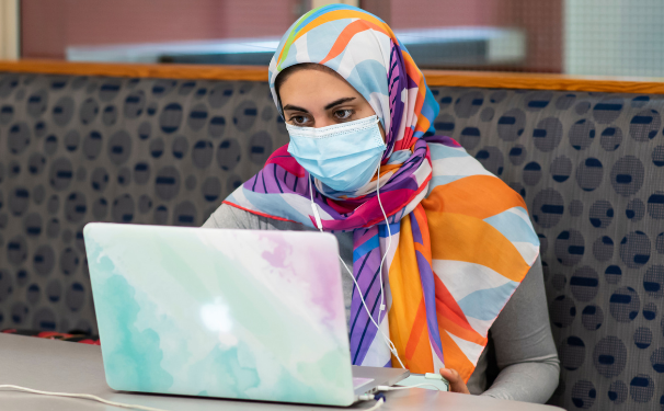 A person studies at a pastel covered laptop. They wear a mask and a hijab.