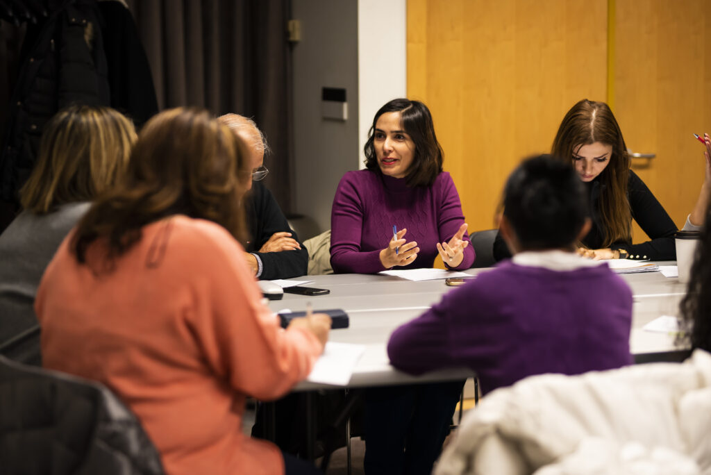 A photo of several people sitting around a table in the library. They are engaged in discussion. A facilitator gestures while speaking to the group.