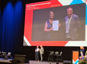 Library Director Stephanie Hall stands on stage holding a paper certificate presented by IFLA Green Library Award coordinator Harri Sahavirta. Behind is a projector screen with "88th IFLA World Library and Information Congress" as the heading.