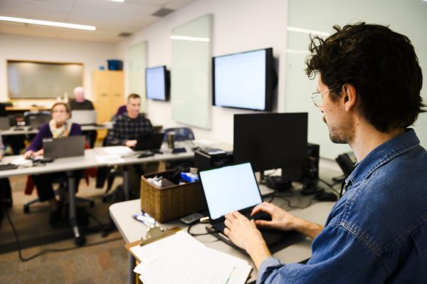 a tech librarian sits at the front of the room, teaching a tech class to a group of adult students using laptop computers