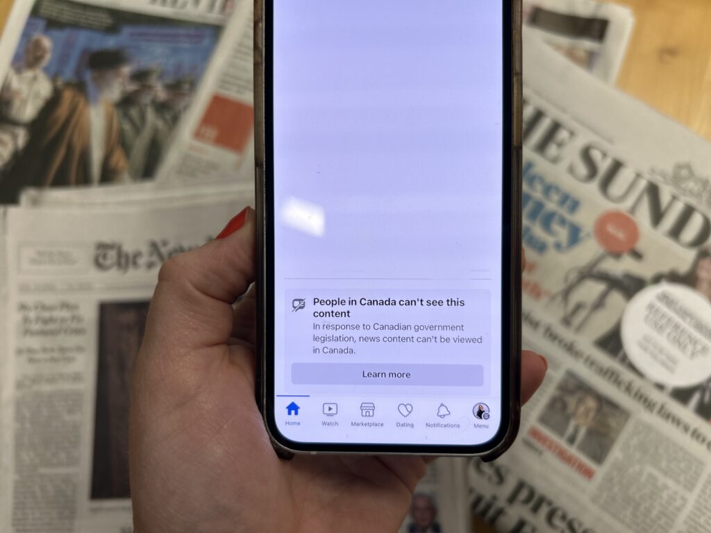 a hand holds a smartphone above a stack of newspapers. the phone screen reads: News content can't be viewed in Canada.