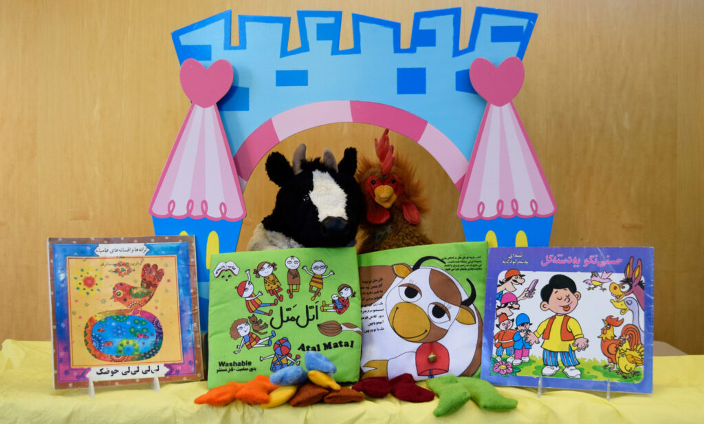 A cow and chicken puppet pose behind picture books written in Farsi
