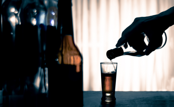 A shadowed hand pours from a bottle into a glass on a table with other bottles off to the side.