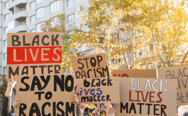 On a city street we see cardboard handpainted signs reading "Black Lives Matter" and "stop Racism"