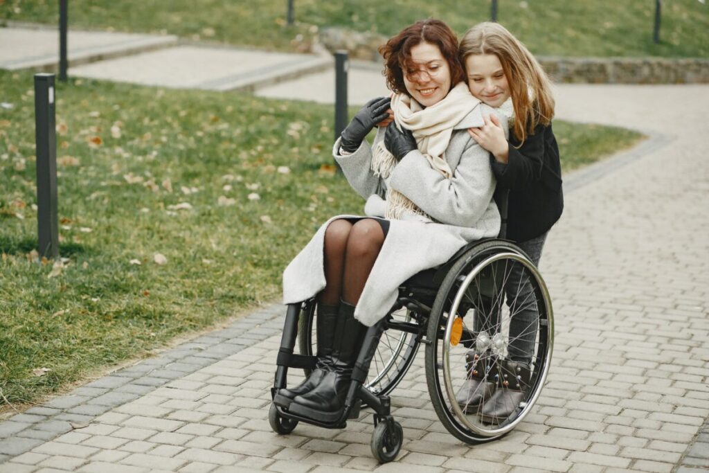 two women--one seated in a wheelchair and one standing behind -- embrace on a sidewalk