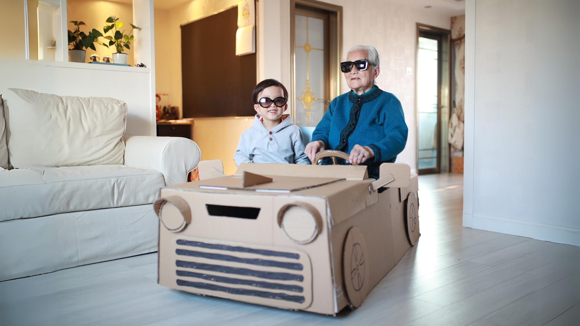An older adult and a young child wear sunglasses while sitting in a cardboard car in a living room.