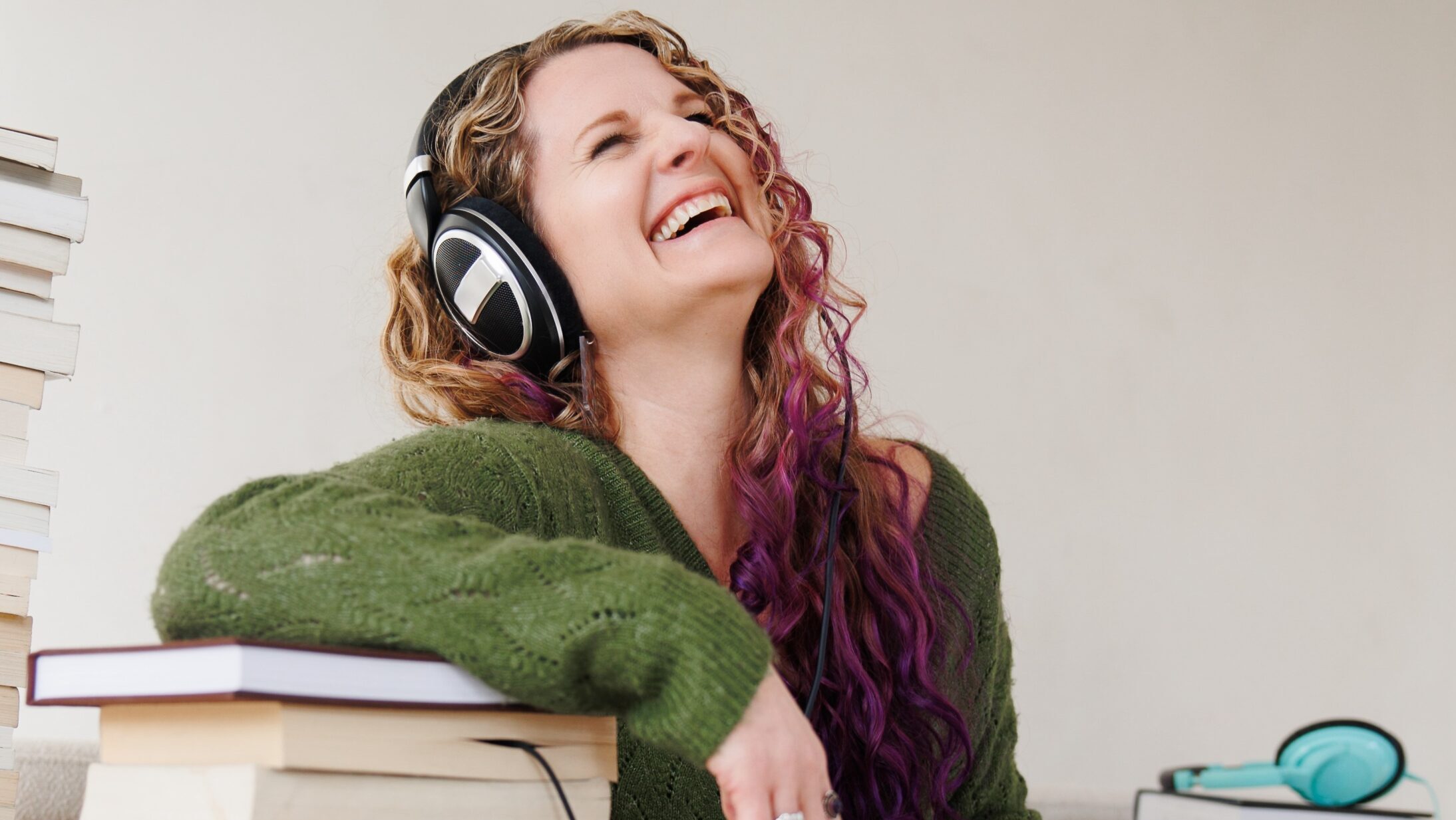 A woman with long curly hair and a green sweater sits among a pile of books listening to an audiobook