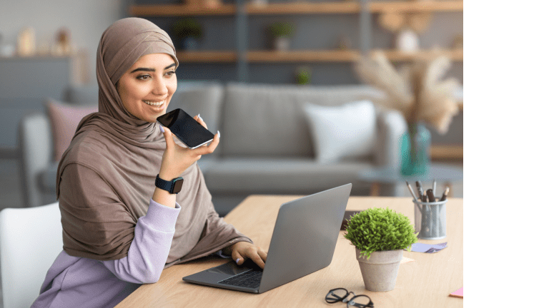 A woman in a hijab sits at a desk while speaking into her smartphone.