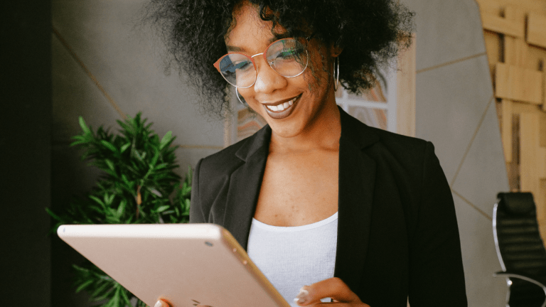 A woman wearing glasses and a blazer smiles at her iPad.