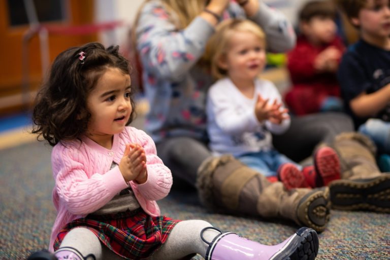 Little kids sit on the floor or in an adult's lap, clapping along to a storytime song.