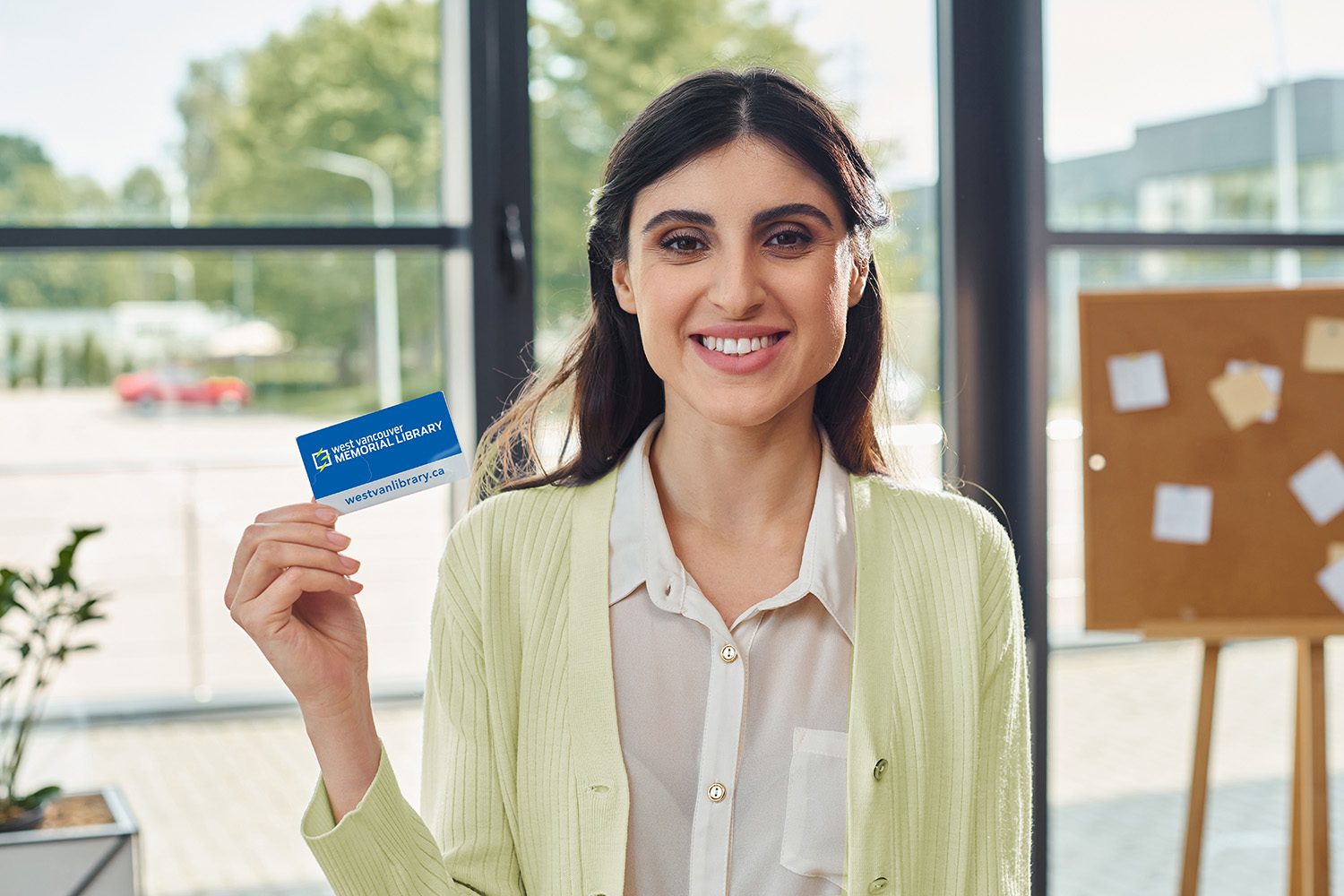 a businesswoman wearing a white collared shirt smiles while holding a WVML library card, with a meeting room sticky note chart in the background.