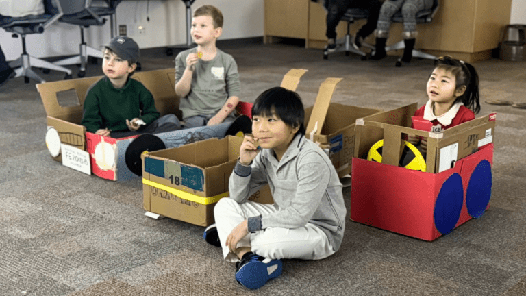 Kids sit in cars made out of cardboard boxes.