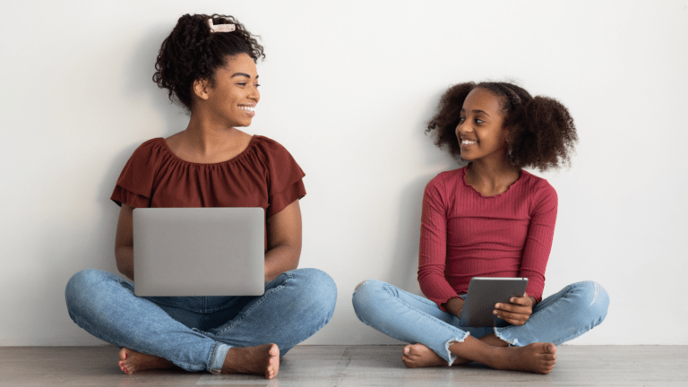 A teen using a laptop and a child using an iPad smile at each other.