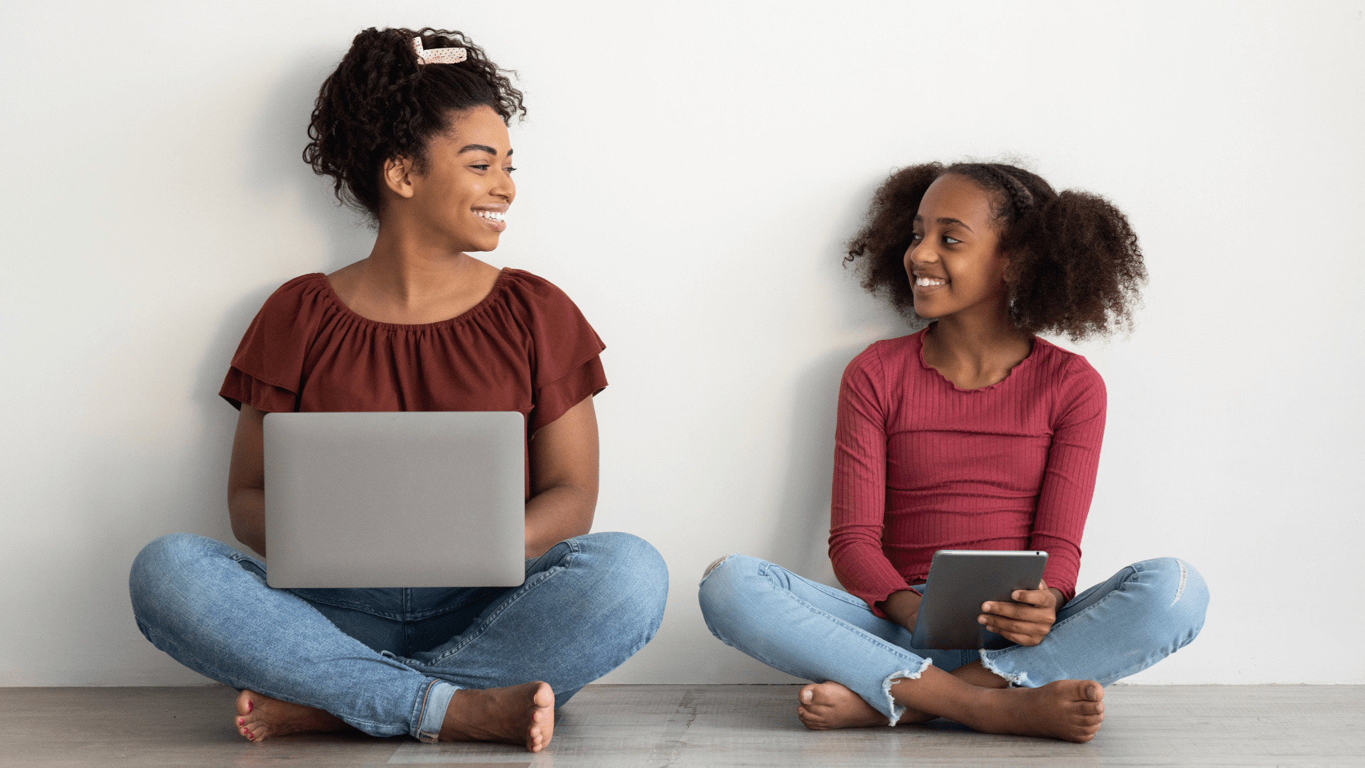 A teen using a laptop and a child using an iPad smile at each other.