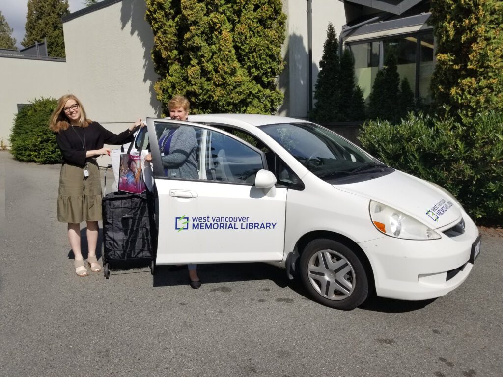 Two Accessible Services staff members stand outside a while car with the Library logo, loading it with bags of books for delivery.