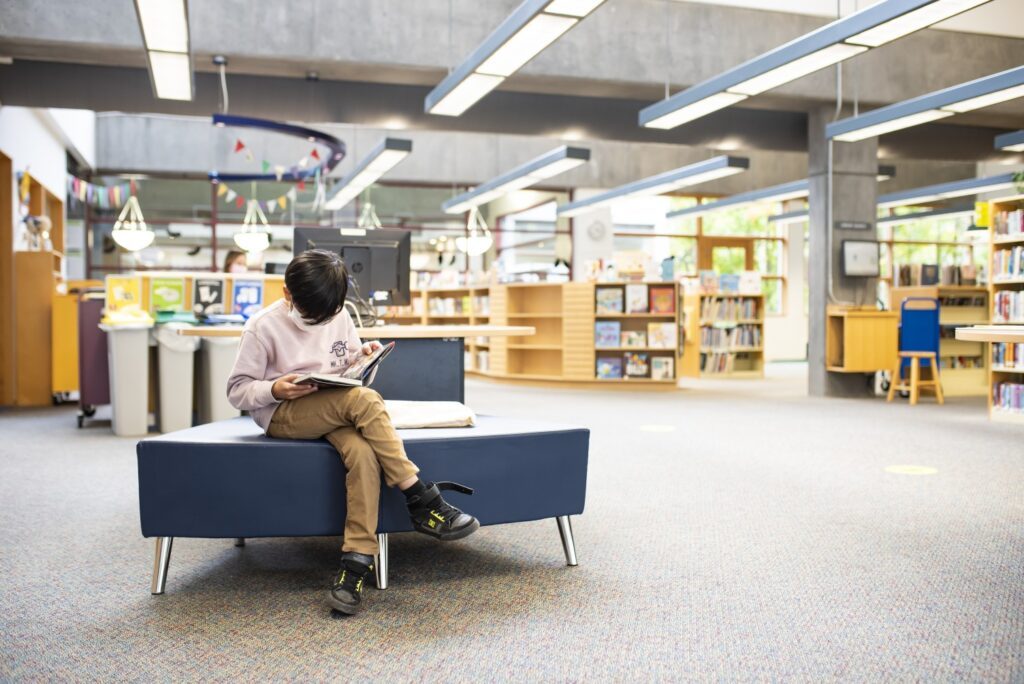 A child sits with his head down reading on a blue bench. In the background are book shelves and displays.