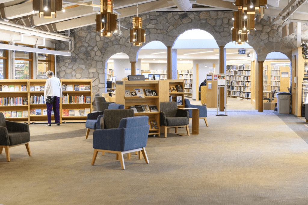 The Main Hall of the library with armchairs and book displays in the middle of the room. Sunlight pours through the windows and an archway to another part of the Library is visible.