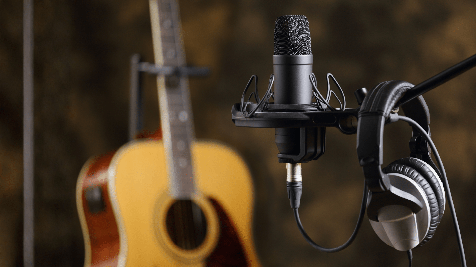 Microphone stand with headphones hanging on it in foreground. Acoustic guitar in background.