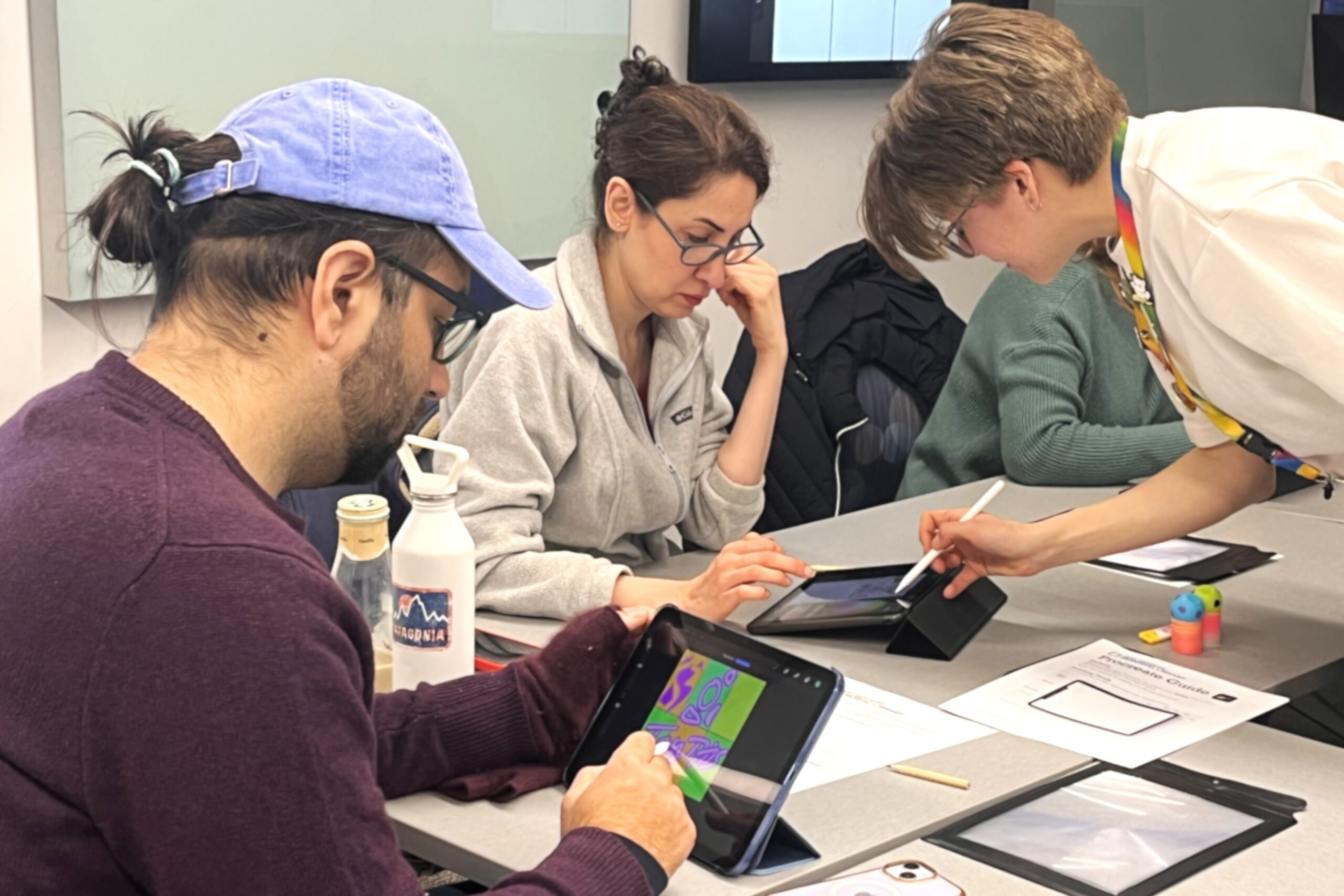 A group of three people sit at a table in a classroom, each drawing on a tablet with a stylus. An instructor leans over one iPad, pointing at the screen with a stylus.