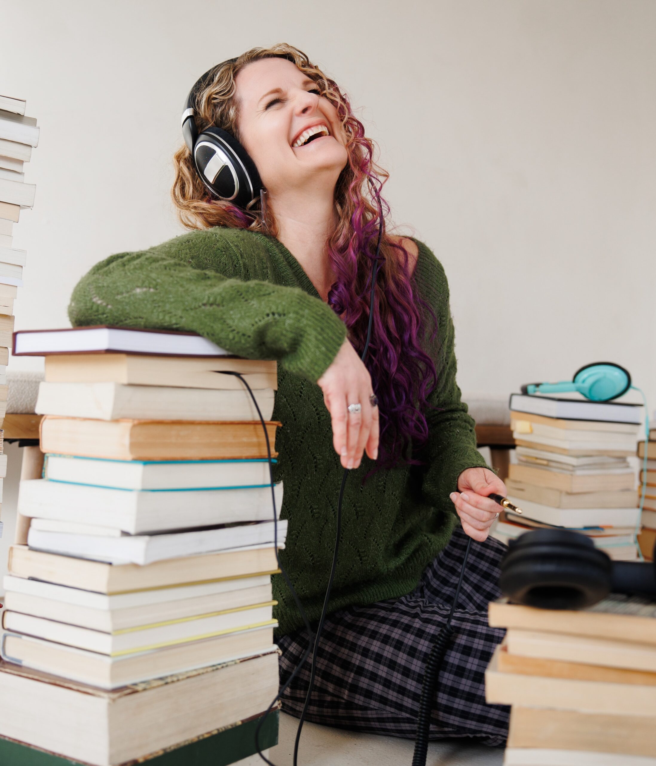 A woman with long reddish hair leans on a pile of books. She is wearing headphones.