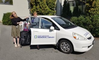 Two Accessible Services staff members stand outside a while car with the Library logo, loading it with bags of books for delivery.
