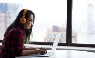 Someone sits by a bright window. wearing orange over-ear headphones and using a laptop.