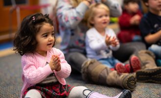 Little kids sit on the floor or in an adult's lap, clapping along to a storytime song.