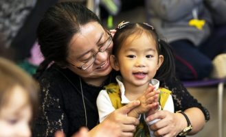 A young girl in a yellow dress with pigtails sits in her mum's lap, clapping and smiling.