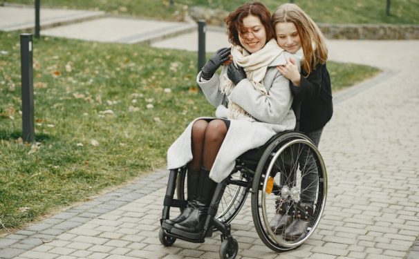 two women--one seated in a wheelchair and one standing behind -- embrace on a sidewalk