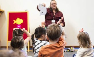 A youth librarian holds up a felt animal in front of a group of kids sitting during storytime.